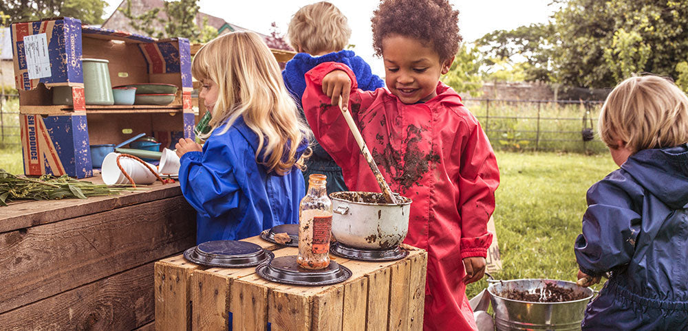 Children in waterproofs playing outdoors with mud kitchen and pots
