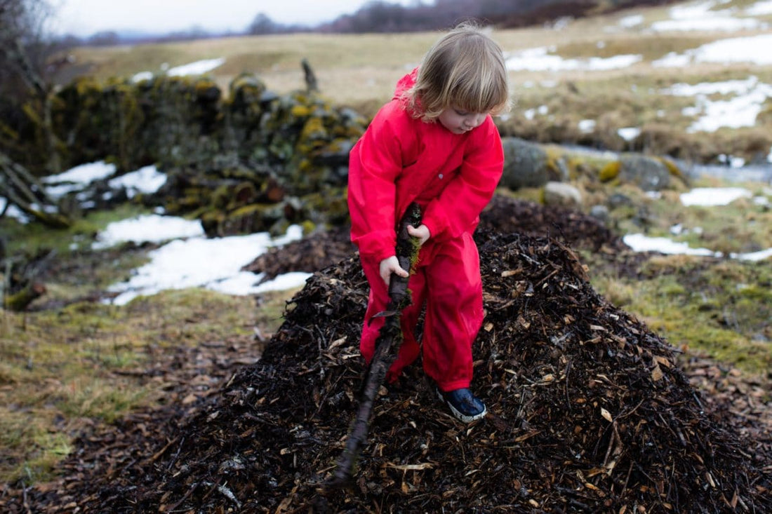 child stood on pile of bark holding a long stick, wearing vibrant red muddy puddles puddlesuit