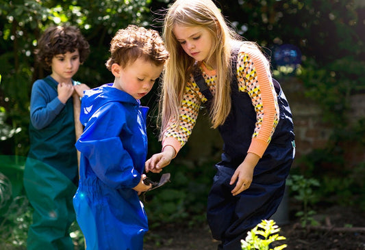 three children gardening using trowel and garden spade, wearing muddy puddles waterproof dungarees and puddlesuit