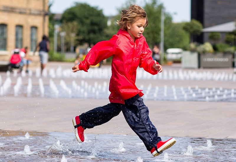 child running through fountains wearing muddy puddles red jacket, waterproof trousers and wellies