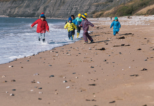 children running along beach wearing vibrant muddy puddles waterproofs and bobble hats