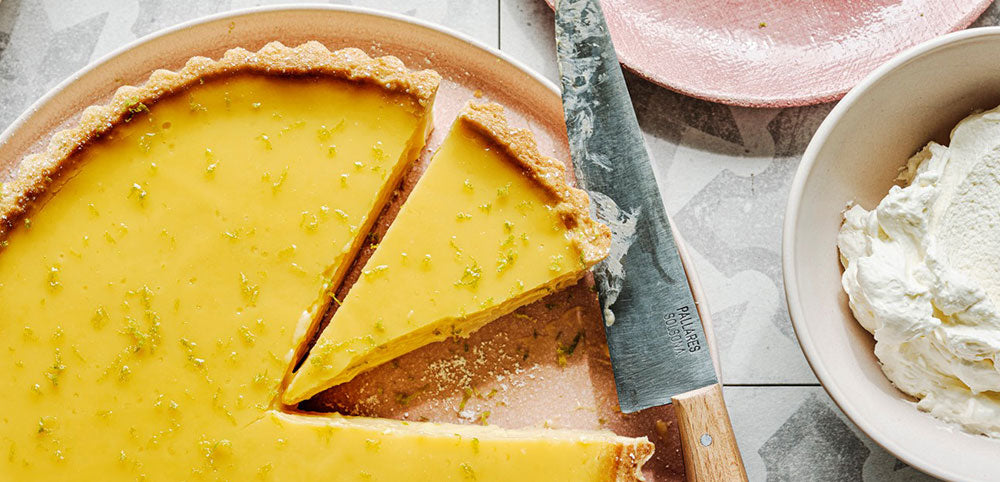 Passionfruit tart with knife, a slice served on a pink plate, and bowl of whipped cream