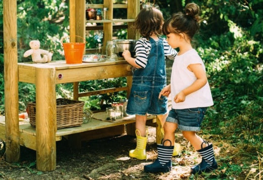 toddler girls playing with a wooden mud kitchen in a shady garden