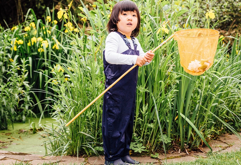 child stood next to pond holding large fishing net and wearing navy waterproof dungarees and wellies