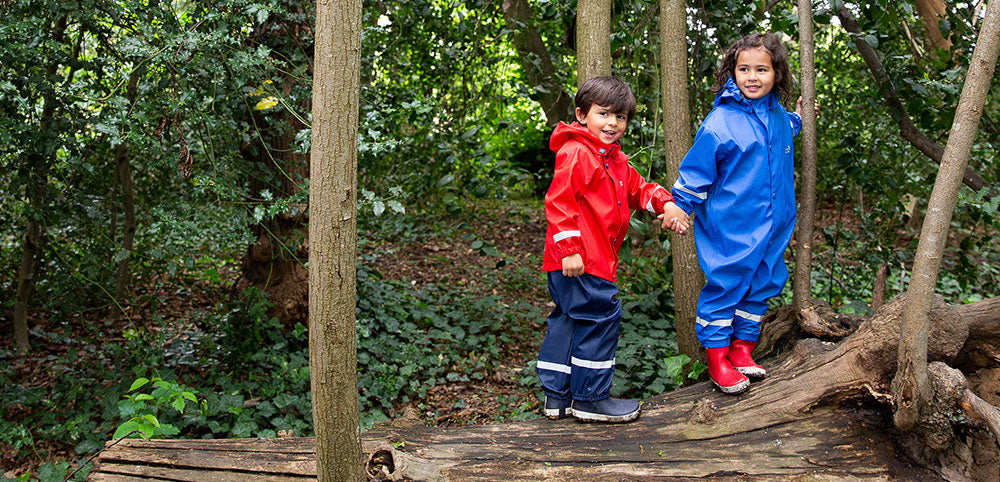 Children in waterproof rain suits exploring a forest, wearing red and blue kids' rainwear.