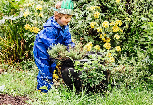 boy in garden planting in large black pot, wearing feather crown and muddy puddles blue waterproof puddlesuit