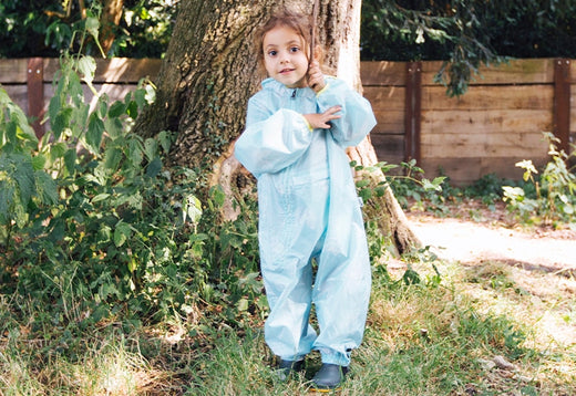 girl stood at base of tree in garden, holding a stick, and wearing light blue muddy puddles puddlesuit