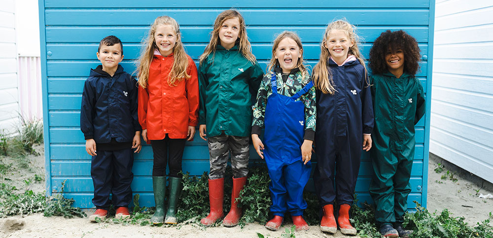 six children stood in front of vibrant blue beach hut wearing muddy puddles originals waterproofs in shades of navy, red, green and blue