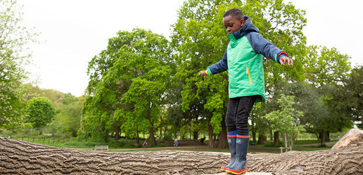 Child in waterproof jacket and rain boots balancing on a log outdoors in a green park