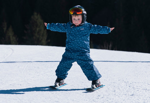 toddler stood up on skis holding arms out for balance, wearing muddy puddles skiwear