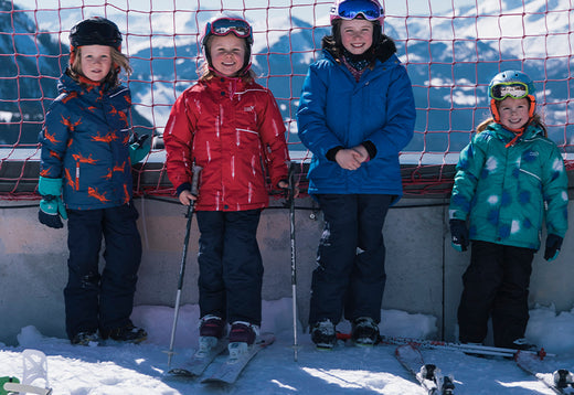 four children wearing skis, stood against fence at snowy ski resort, wearing muddy puddles skiwear