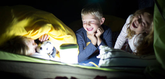 Three children laughing and talking in a tent at night, illuminated by a lantern, cozy bedding visible.