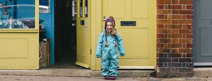 girl stood in front of yellow door, wearing green muddy puddles puddlesuit and trapper hat