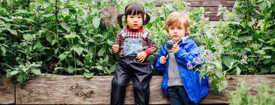 boy and girl in garden wearing muddy puddles waterproofs. Boy is holding a magnifying glass.