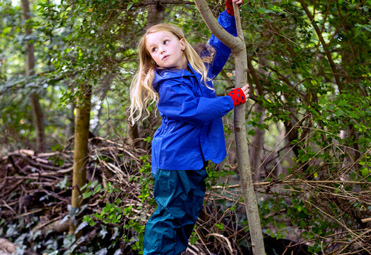 blonde girl in woodland holding tree branch, wearing vibrant blue muddy puddles waterproof jacket and navy waterproof trousers