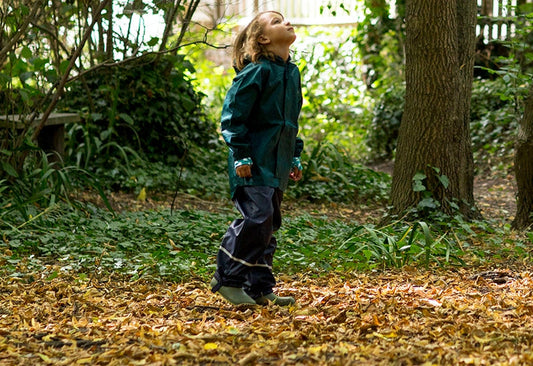 child stood in woodland setting, looking up to treetops, wearing green and navy muddy puddles waterproofs