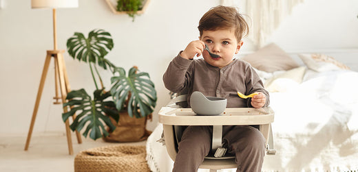 Young child in grey outfit eating with spoon in high chair, cozy indoor setting
