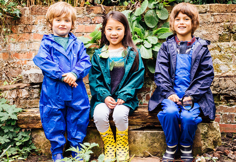 children sitting on bench in front of brick wall, wearing colourful muddy puddles puddlesuit, waterproof jacket, and waterproof dungarees