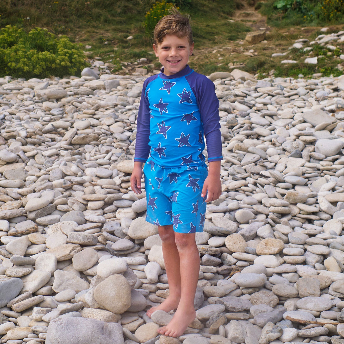 A boy stands on a pebbled beach in a 'UV Protective Rash Vest Blue Starfish', smiling at the camera.