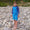 A boy stands on a pebbled beach in a 'UV Protective Rash Vest Blue Starfish', smiling at the camera.