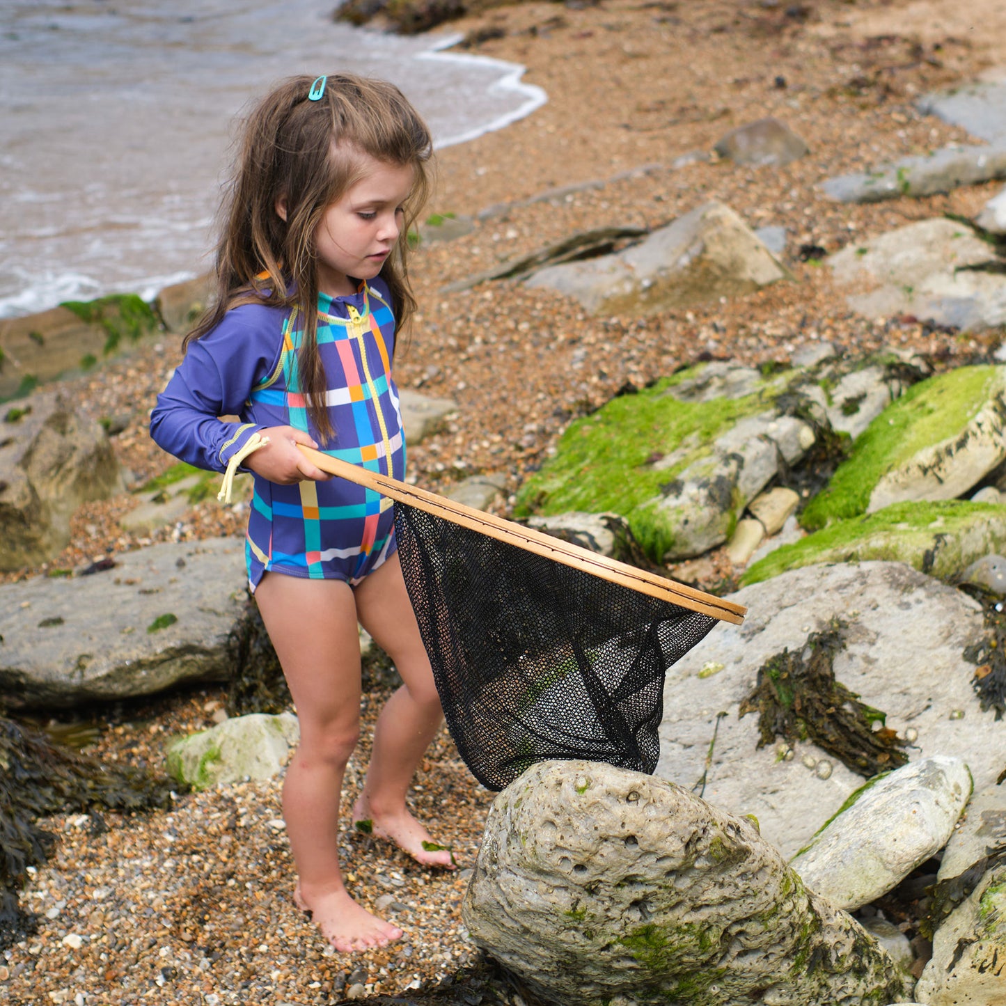 A young child in a UV Protective Long Sleeve Swimsuit Multi Check is on the beach holding a net.