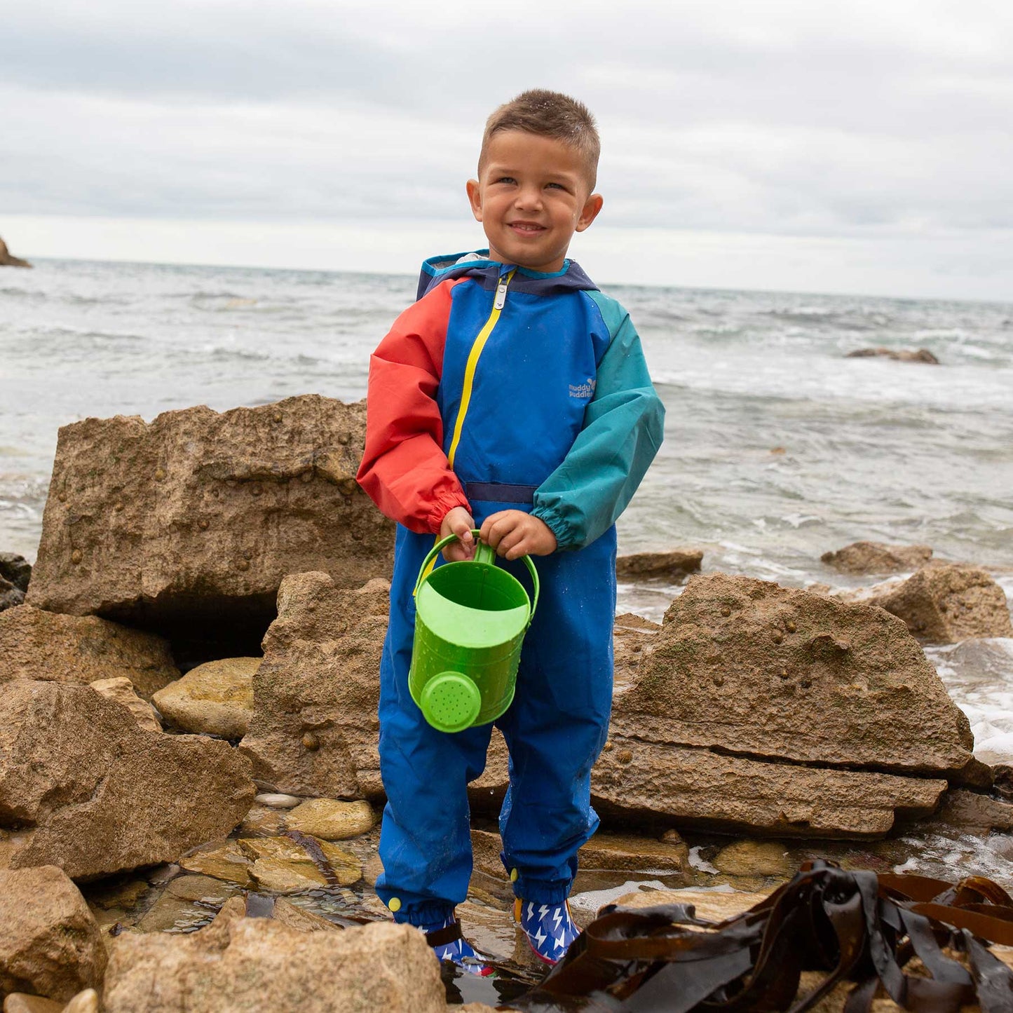 Young boy wearing the EcoLight Lightweight Waterproof Puddle Suit Blue Colourblock stands among rocks, holding a green bucket, showcasing the suit's durable, waterproof design ideal for rainy adventures.