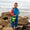 Young boy wearing the EcoLight Lightweight Waterproof Puddle Suit Blue Colourblock stands among rocks, holding a green bucket, showcasing the suit's durable, waterproof design ideal for rainy adventures.