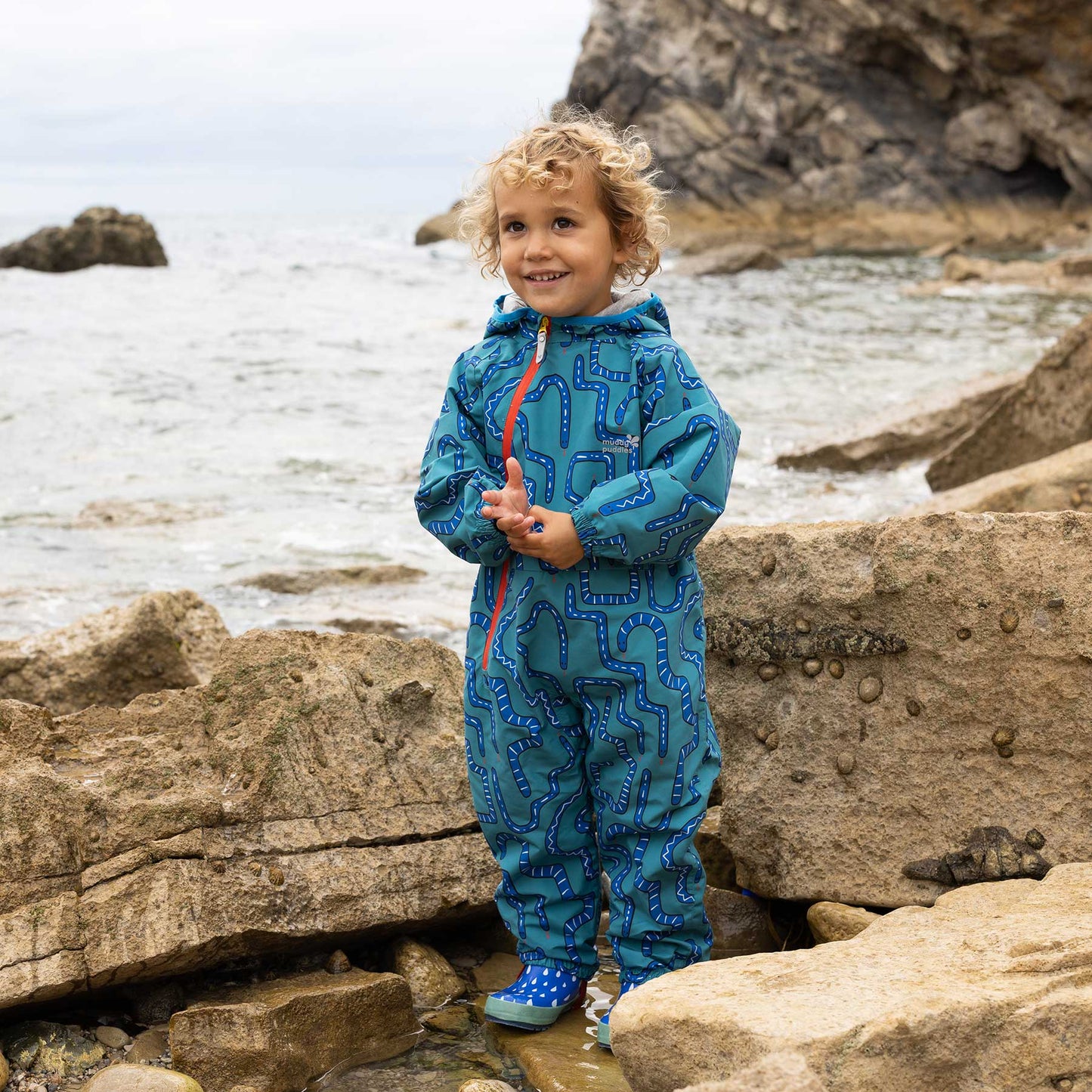 Child wearing EcoLight Lightweight Waterproof Puddle Suit Green Snake on rocky shoreline, showcasing its durable, waterproof design ideal for adventurous, outdoor play in wet conditions.