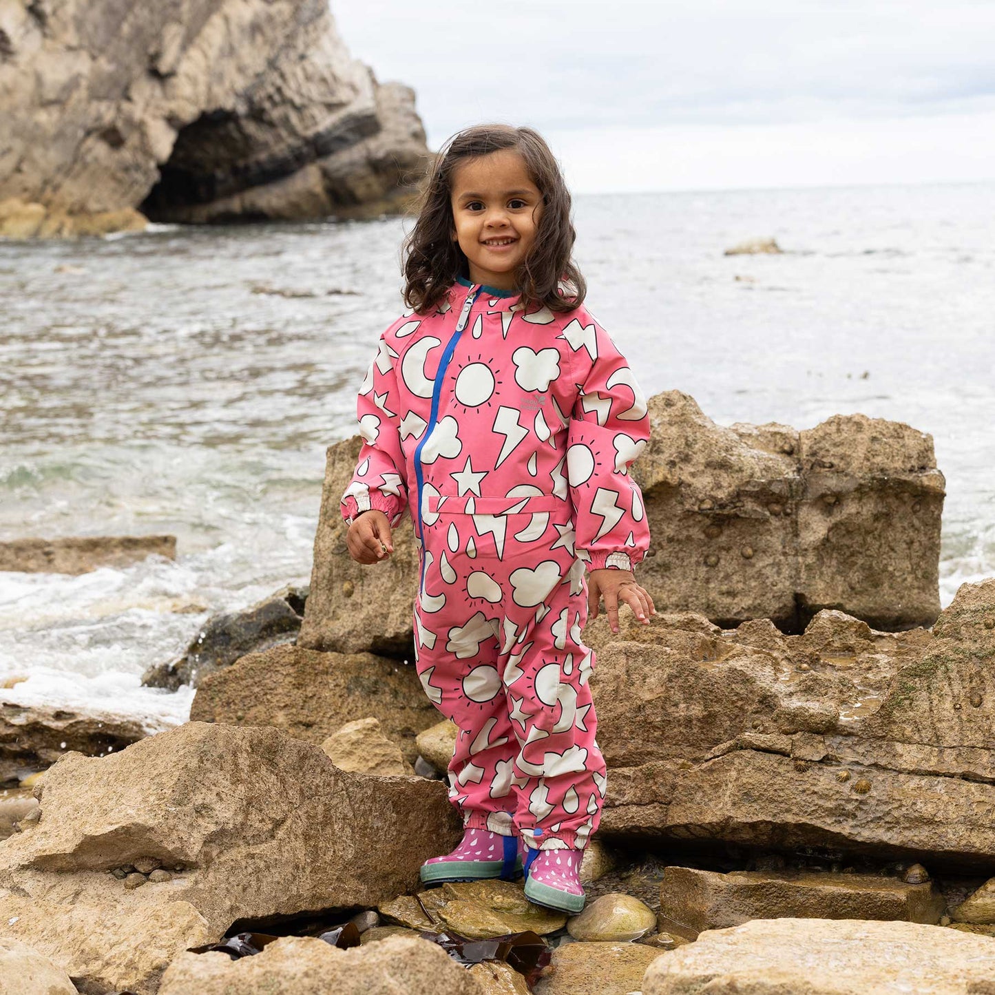 Preschool girl wearing EcoLight Lightweight Waterproof Puddle Suit Pink Weather, exploring a rocky shoreline. The durable, waterproof suit features playful weather-themed prints and cozy lining.