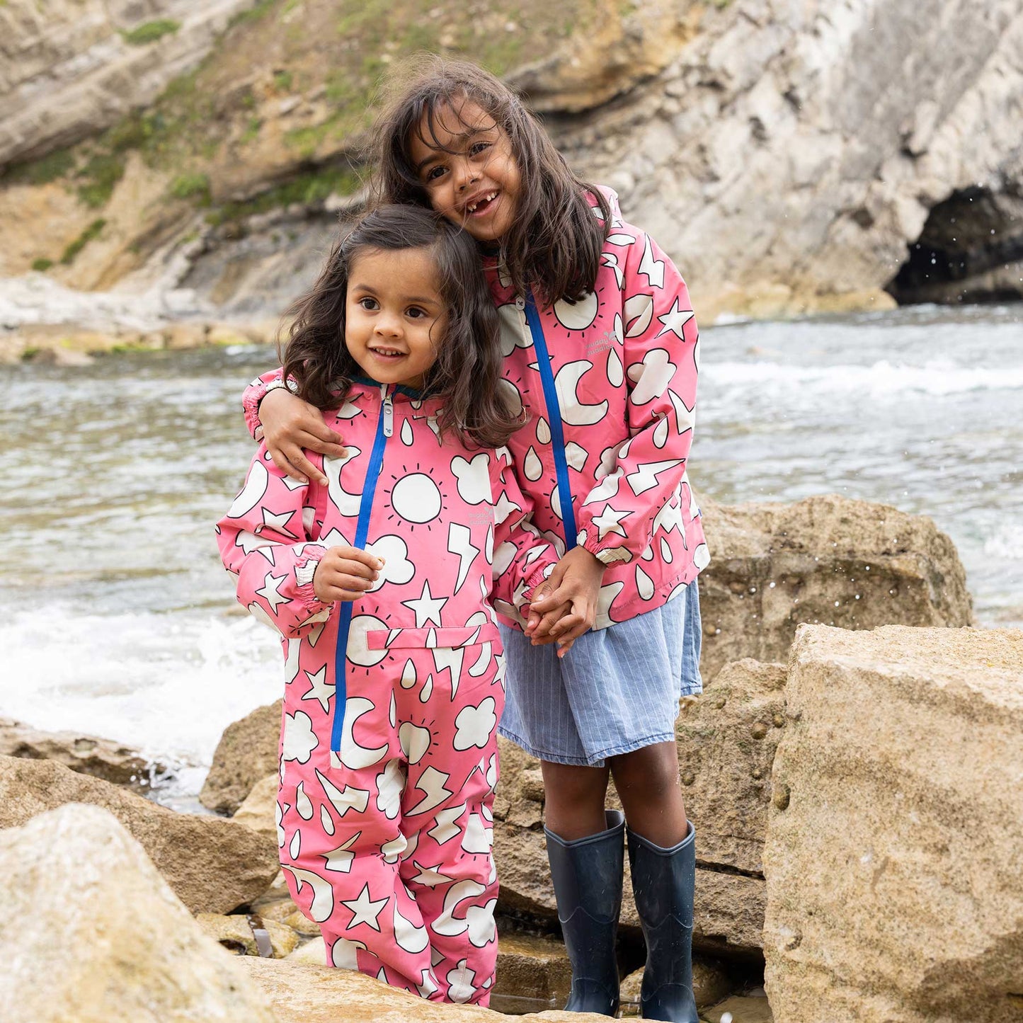 Two girls in EcoLight Lightweight Waterproof Puddle Suits smile by a stream, showcasing the durable, weather-themed print and waterproof design for outdoor adventures.