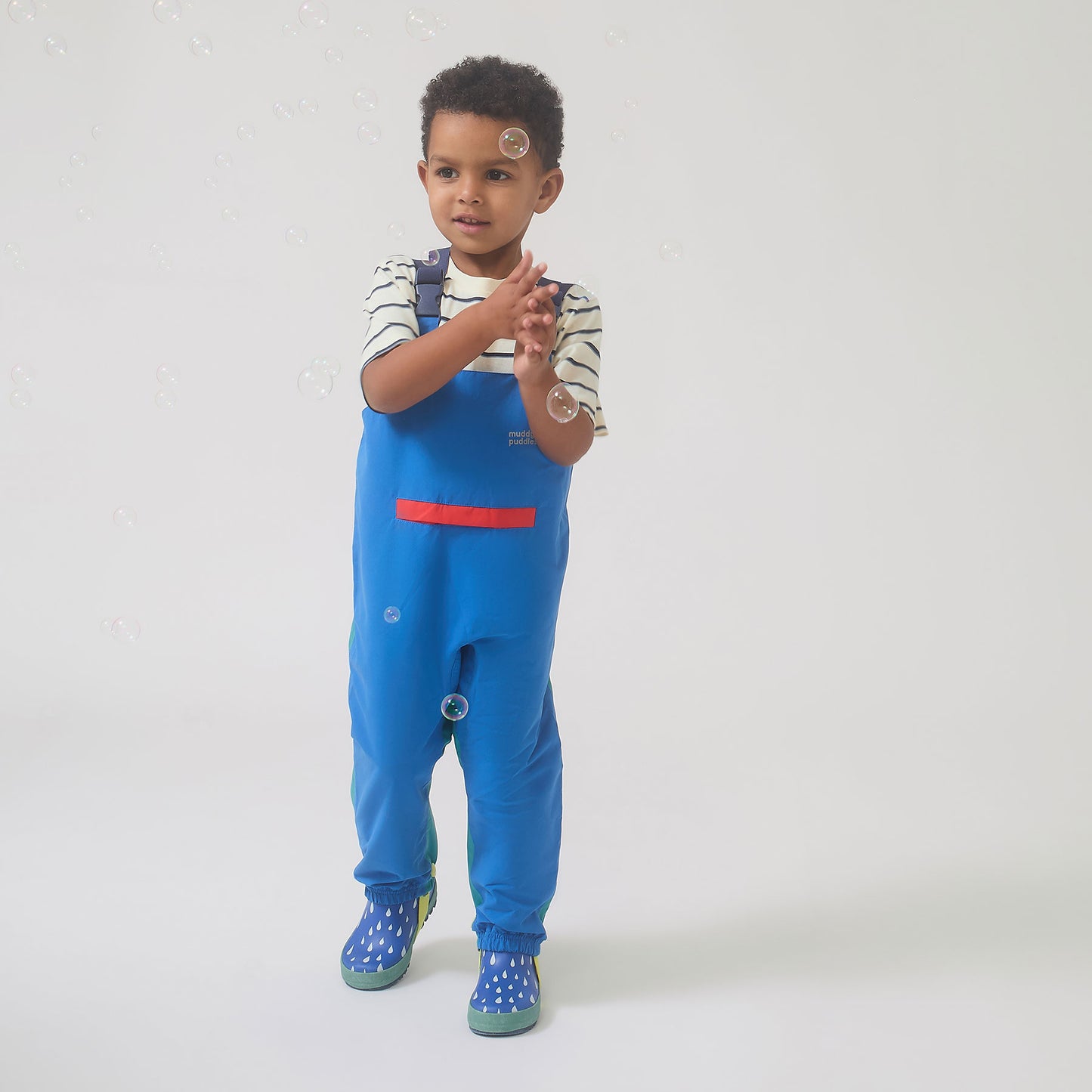 Young boy in EcoLight Waterproof Dungarees Blue Colourblock, playing with bubbles. He's in durable, breathable overalls with adjustable straps, ideal for wet-weather adventures.