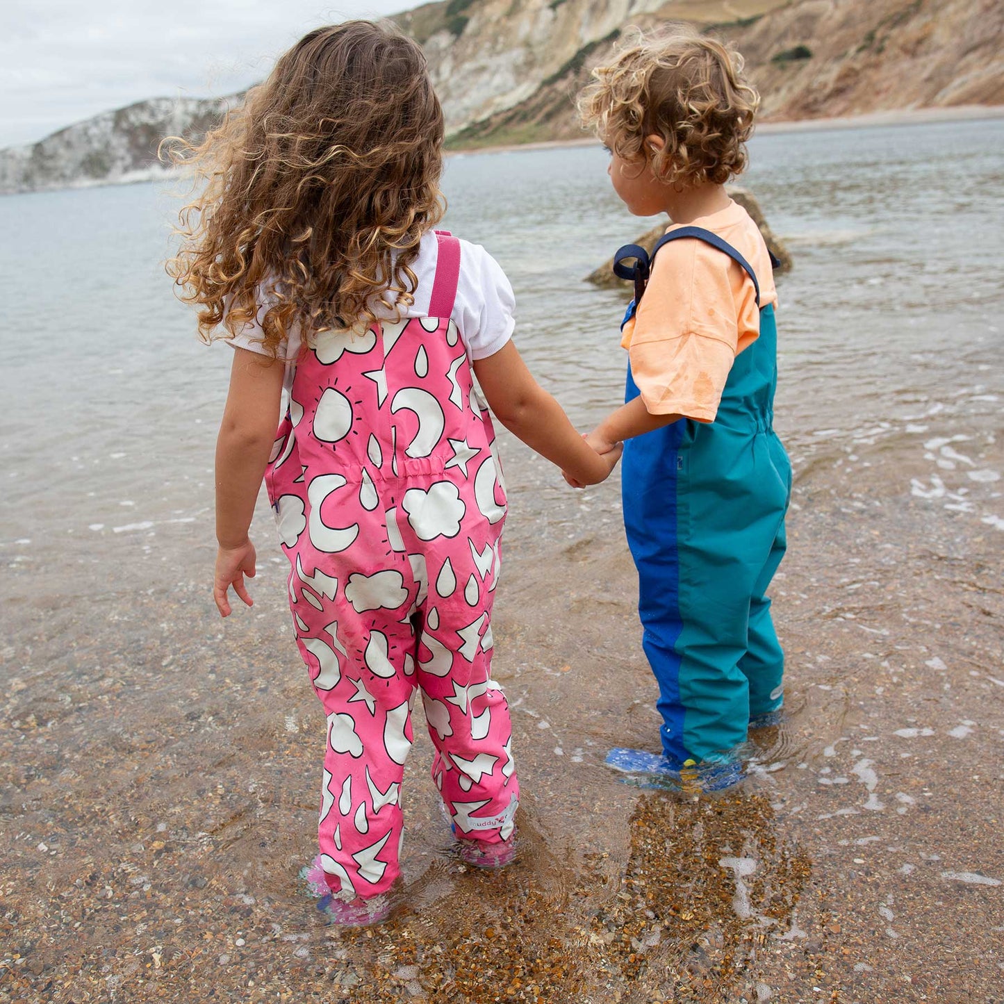 Two children wearing EcoLight Waterproof Dungarees look out to sea, showcasing durable, waterproof outdoor clothing ideal for active play in wet conditions.