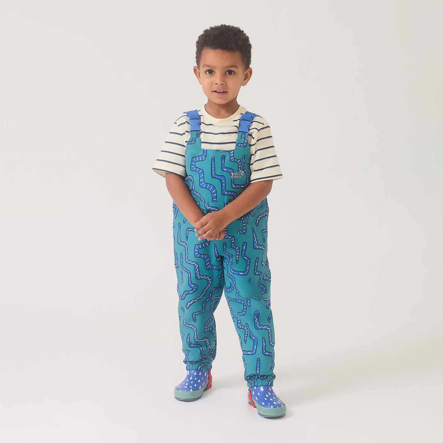 Young boy wearing EcoLight Waterproof Dungarees Green Snake, featuring playful snake-themed print, teal with dark-blue lines, paired with matching rain boots for rainy-day play.