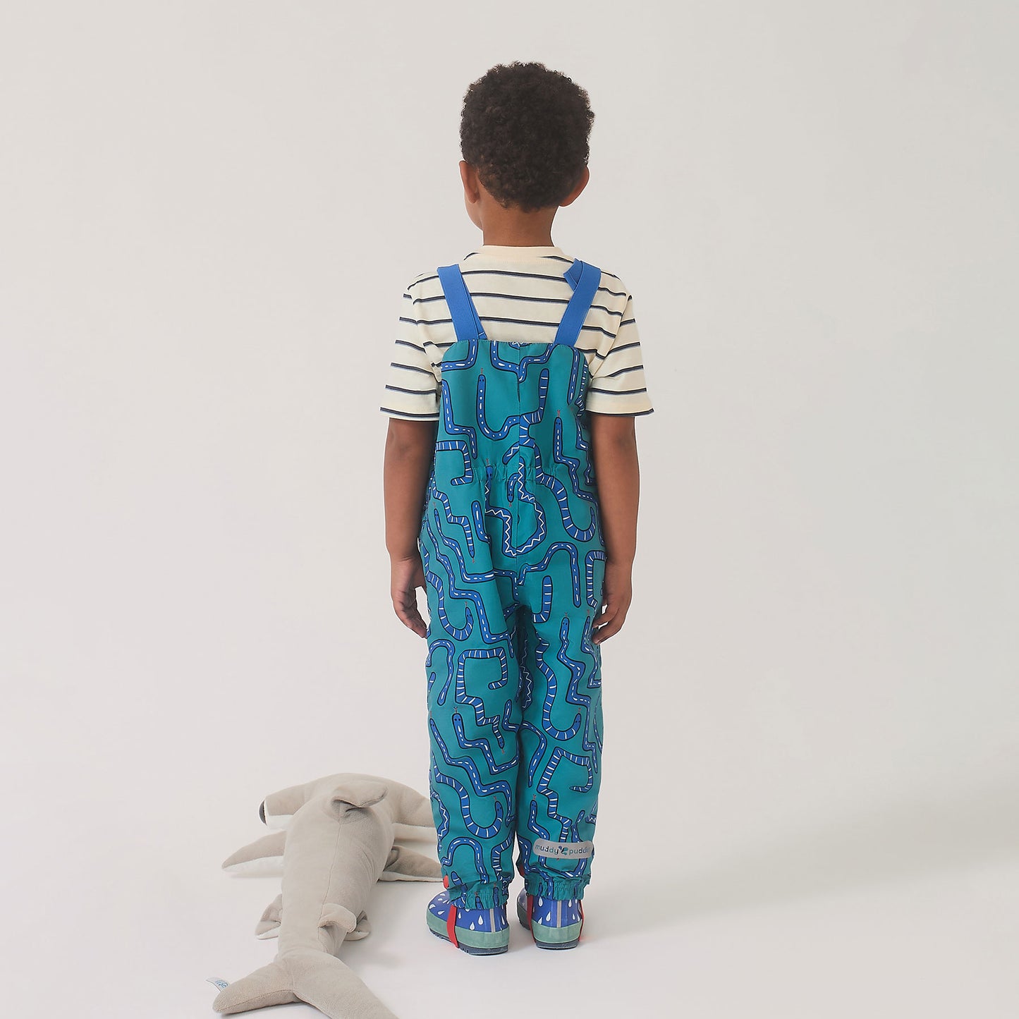 Child wearing EcoLight Waterproof Dungarees Green Snake with adjustable straps, playful snake print, and matching boots, standing beside a gray shark plush toy.