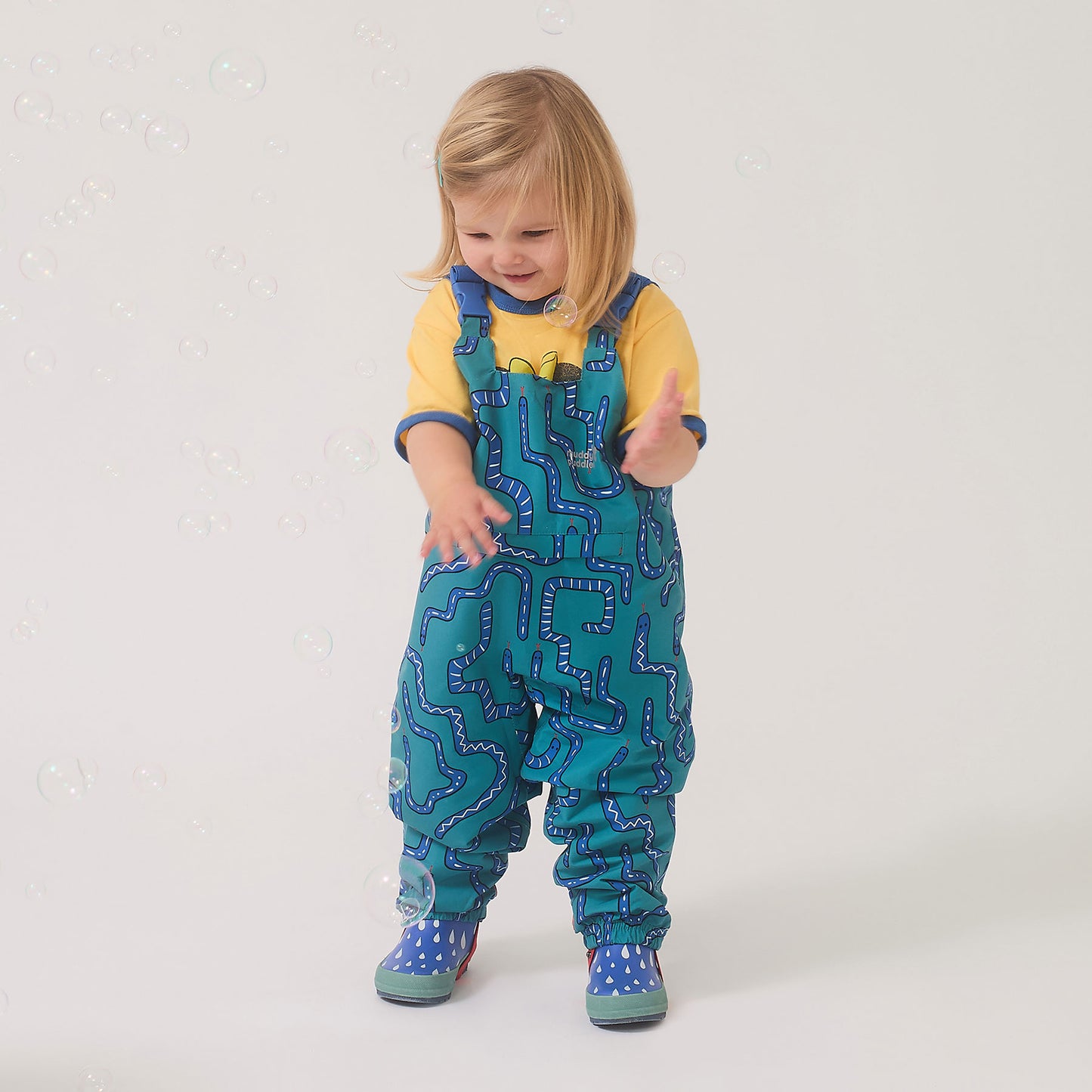 Toddler wearing EcoLight Waterproof Dungarees Green Snake, enjoying bubbles. Features include durable, fluorine-free waterproof fabric, adjustable fit, and playful snake-themed print.