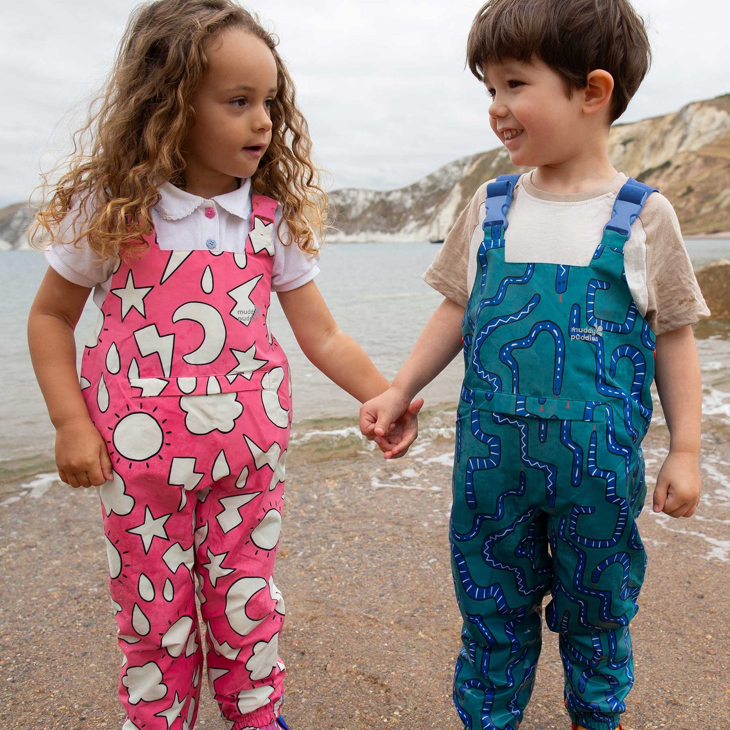Two children wearing EcoLight Waterproof Dungarees Green Snake stand on a beach. The durable, waterproof overalls feature playful patterns, ideal for adventurous, weather-resistant play.