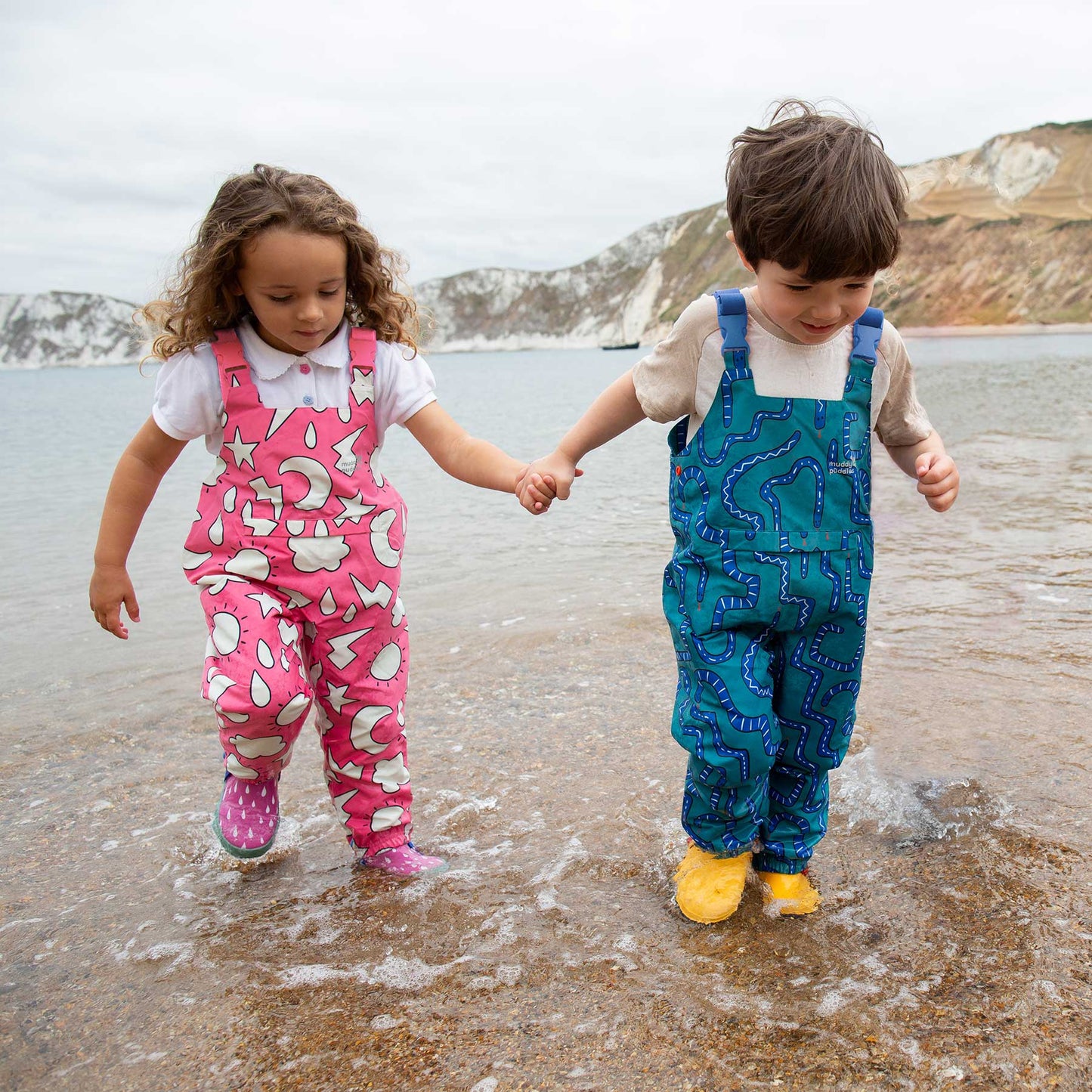 Children in EcoLight Waterproof Dungarees Pink Weather wade through seawater, showcasing durable, waterproof protection with adjustable straps and eco-friendly design from Muddy Puddles.