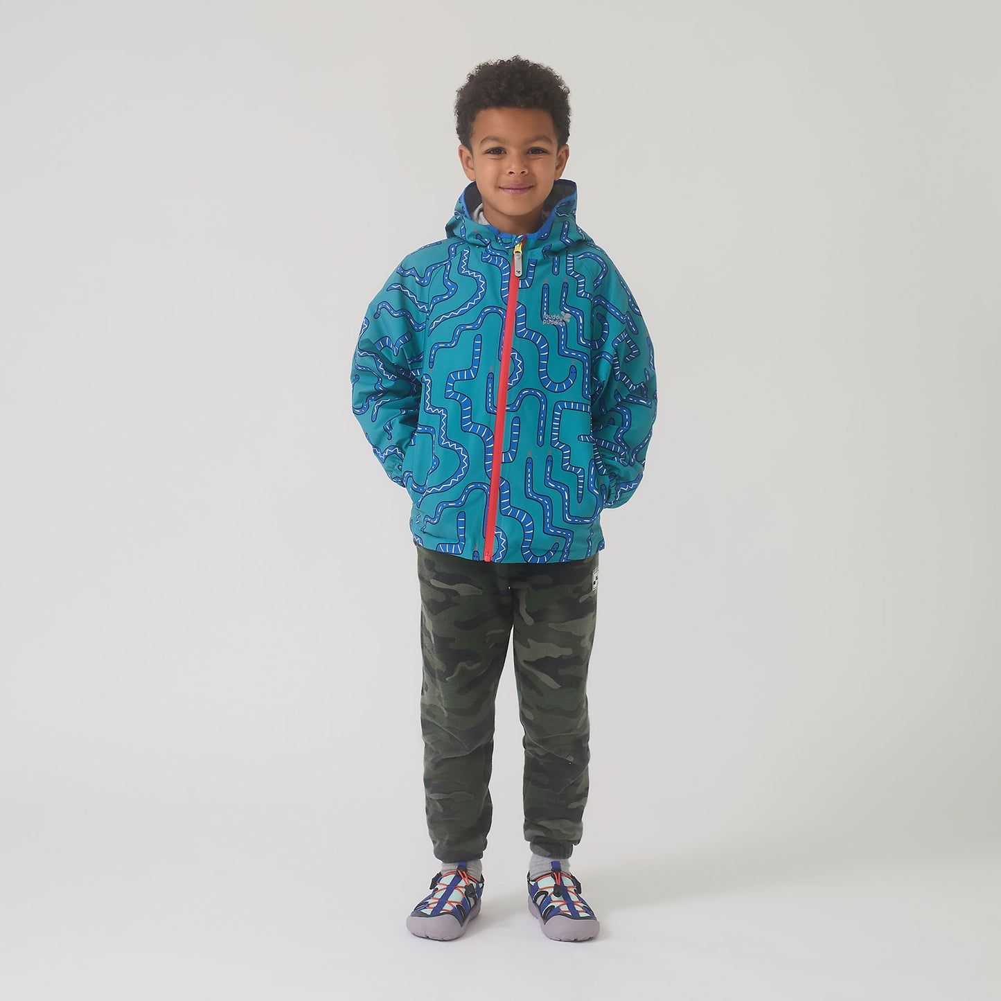 Young boy wearing the EcoLight Lightweight Waterproof Jacket Green Snake, featuring a maze-like pattern, red zipper, and Velcro pockets, paired with camouflage joggers and sporty sandals.