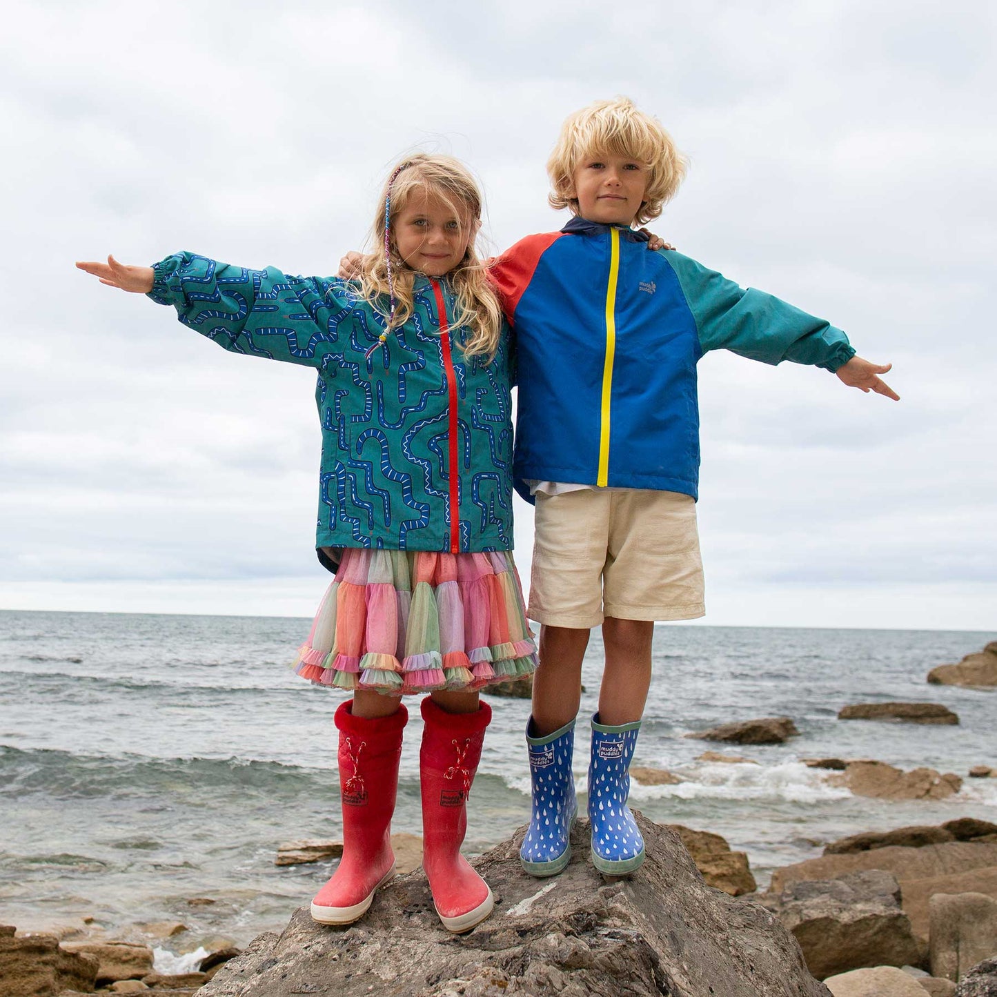 Children wearing EcoLight Lightweight Waterproof Jacket Green Snake balancing on rocky shore, showcasing its durable, waterproof design ideal for outdoor play in wet conditions.