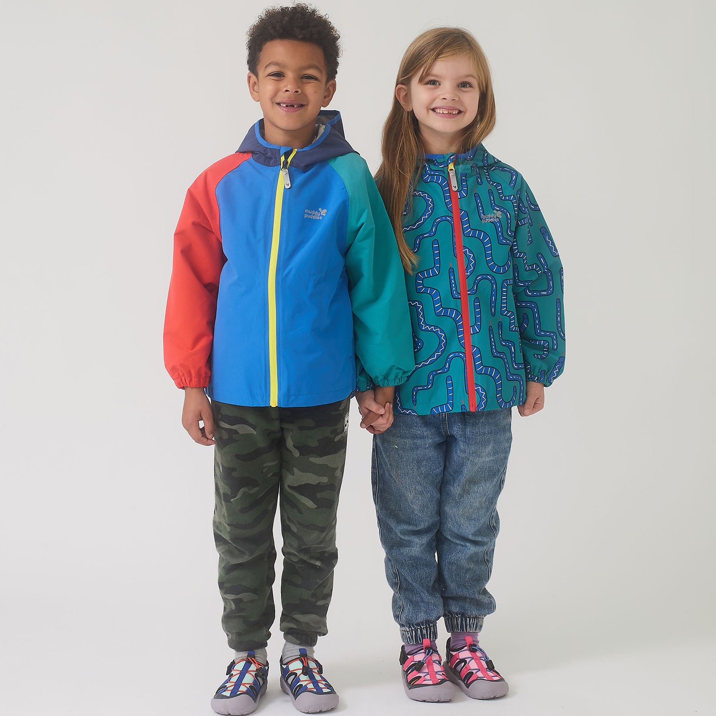 Two children wearing Muddy Puddles EcoLight Lightweight Waterproof Jacket Green Snake, smiling and holding hands, showcasing durable, waterproof outerwear ideal for adventurous play in wet weather.