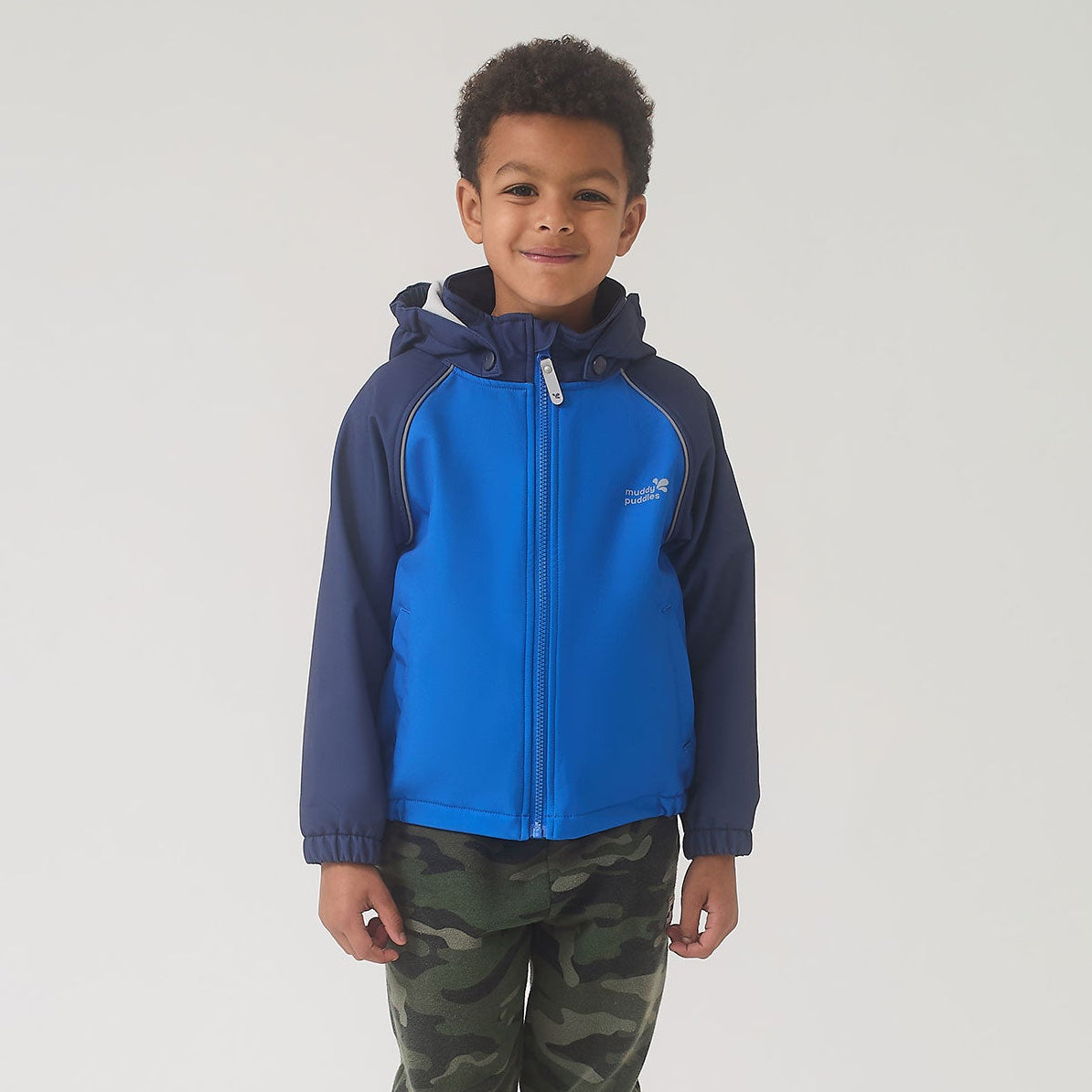 Boy wearing the Waterproof Softshell Jacket Blue, featuring a zip-front, chin guard, and reflective details, paired with camouflage pants and yellow rain boots.