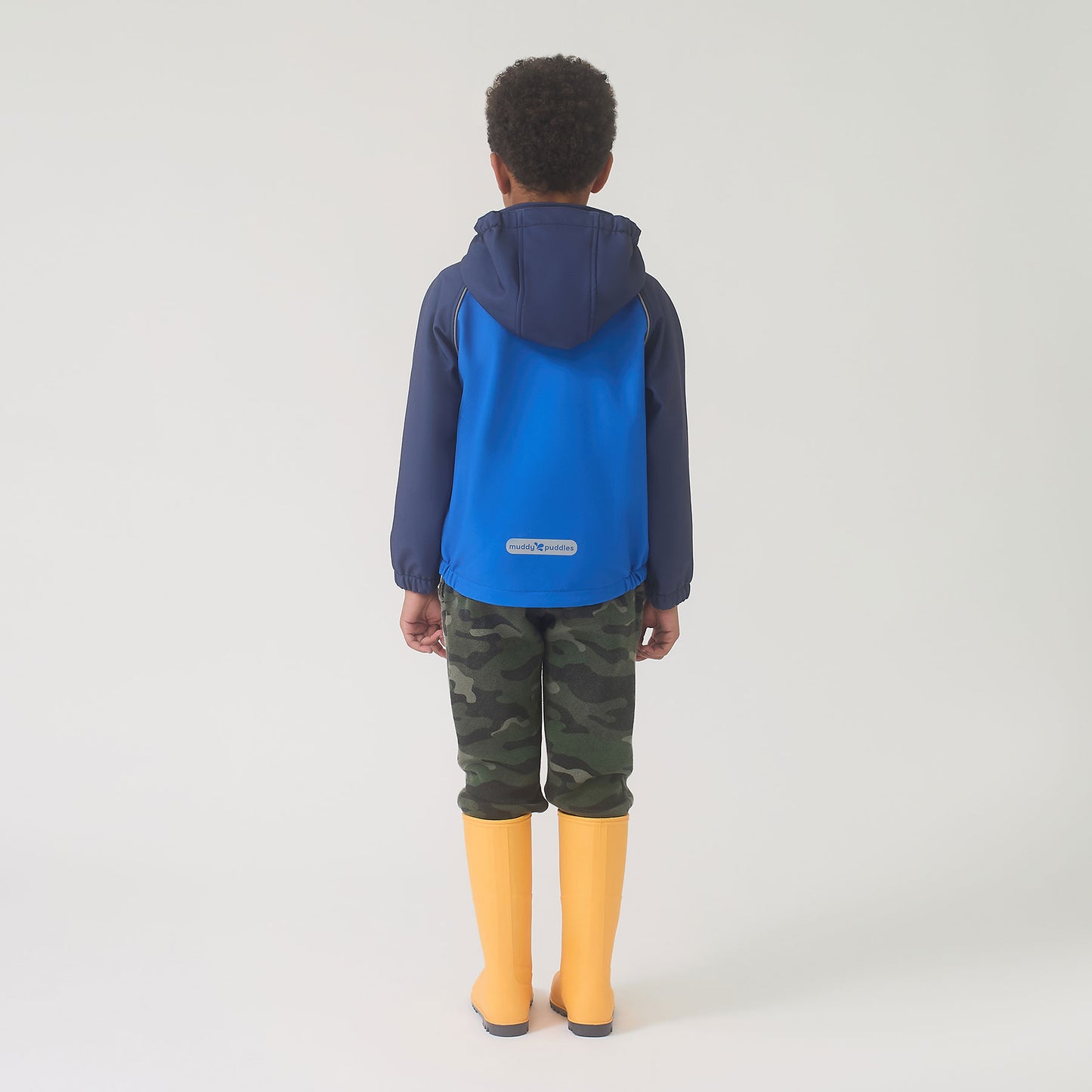 Child wearing the Waterproof Softshell Jacket Blue, featuring a hood, elasticated cuffs, and reflective details, paired with camouflage pants and yellow rain boots, standing ready for rainy adventures.