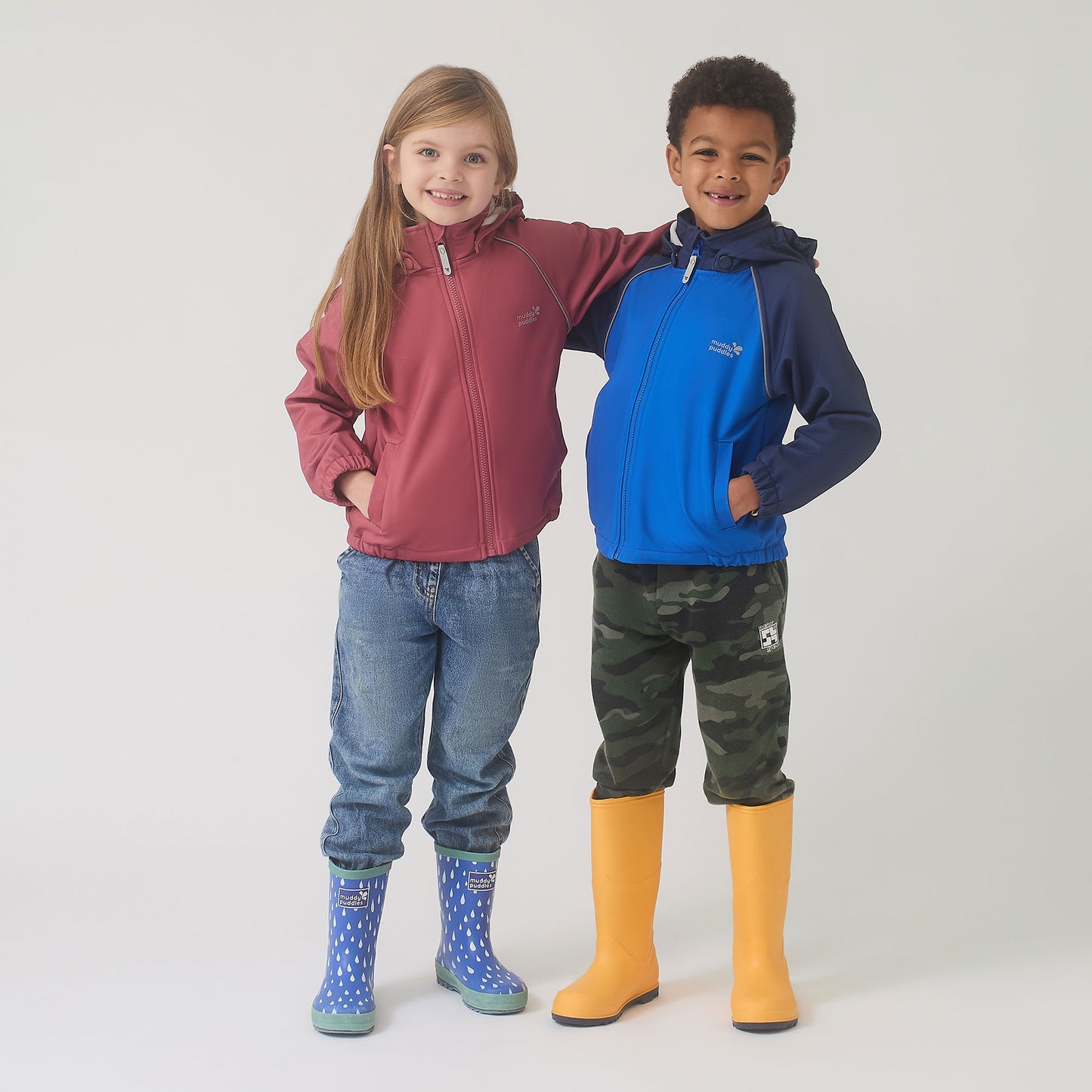 Two children wearing Muddy Puddles' durable Waterproof Softshell Jacket Pink, showcasing its protective features for outdoor adventures in unpredictable weather.