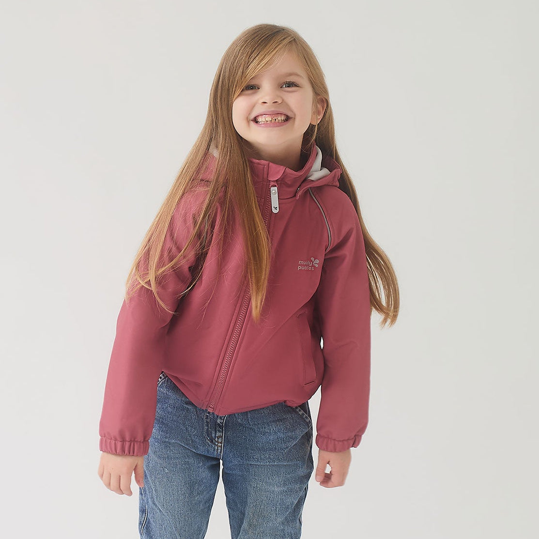 Young girl in a waterproof softshell jacket pink, cuffed jeans, and rain boots. The jacket features a hood, chin guard, and reflective details for durability and outdoor play.