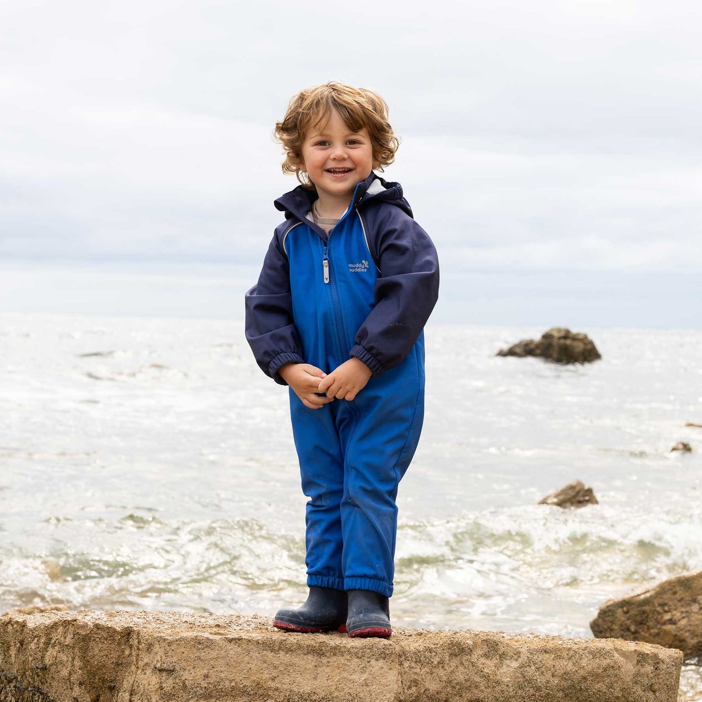 Toddler stands in Waterproof Softshell Puddle Suit Blue on a shoreline rock, showcasing durable, waterproof protection for adventurous outdoor play in challenging weather.
