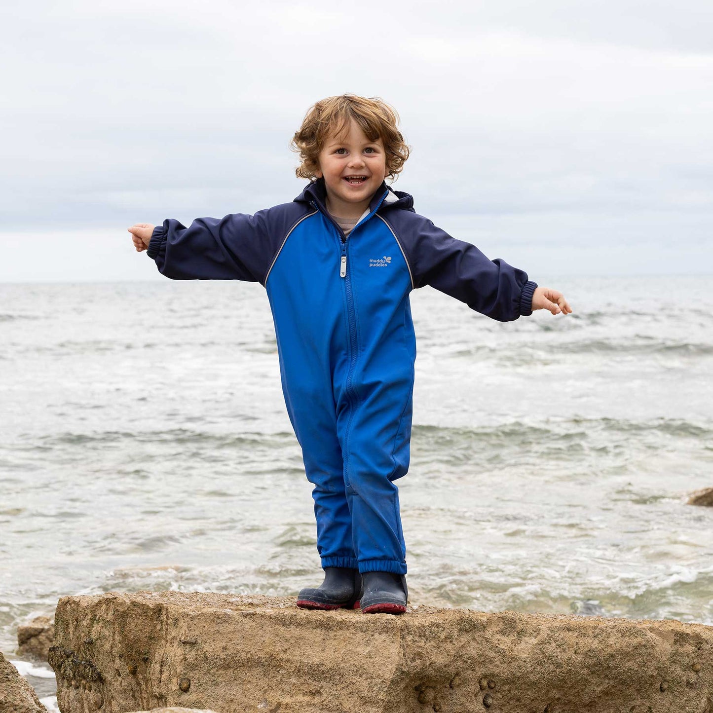 Toddler balances on rock in a Waterproof Softshell Puddle Suit Blue, showcasing its durable, waterproof design ideal for adventurous play in wet conditions.