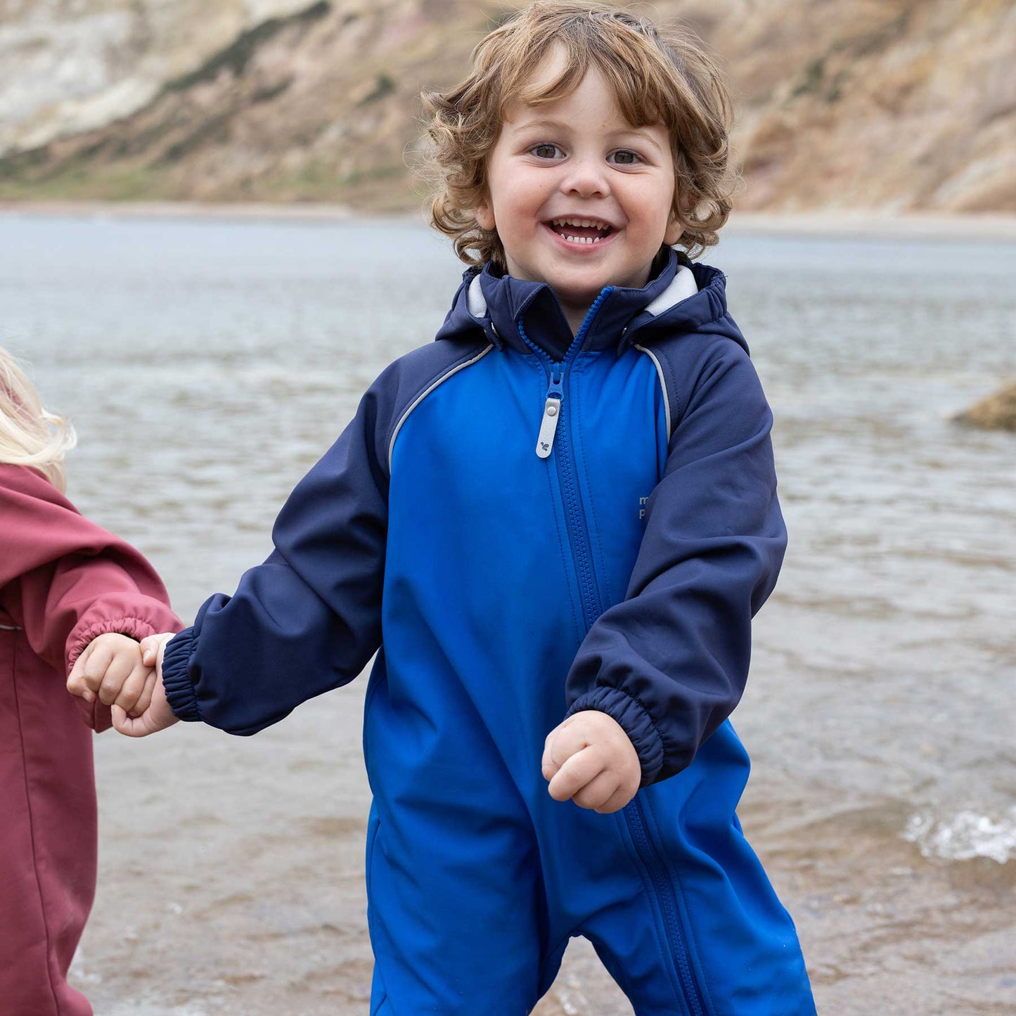 Toddler smiling by water in a durable Waterproof Softshell Puddle Suit Blue, holding hands with another child. Ideal for adventurous, weatherproof play.