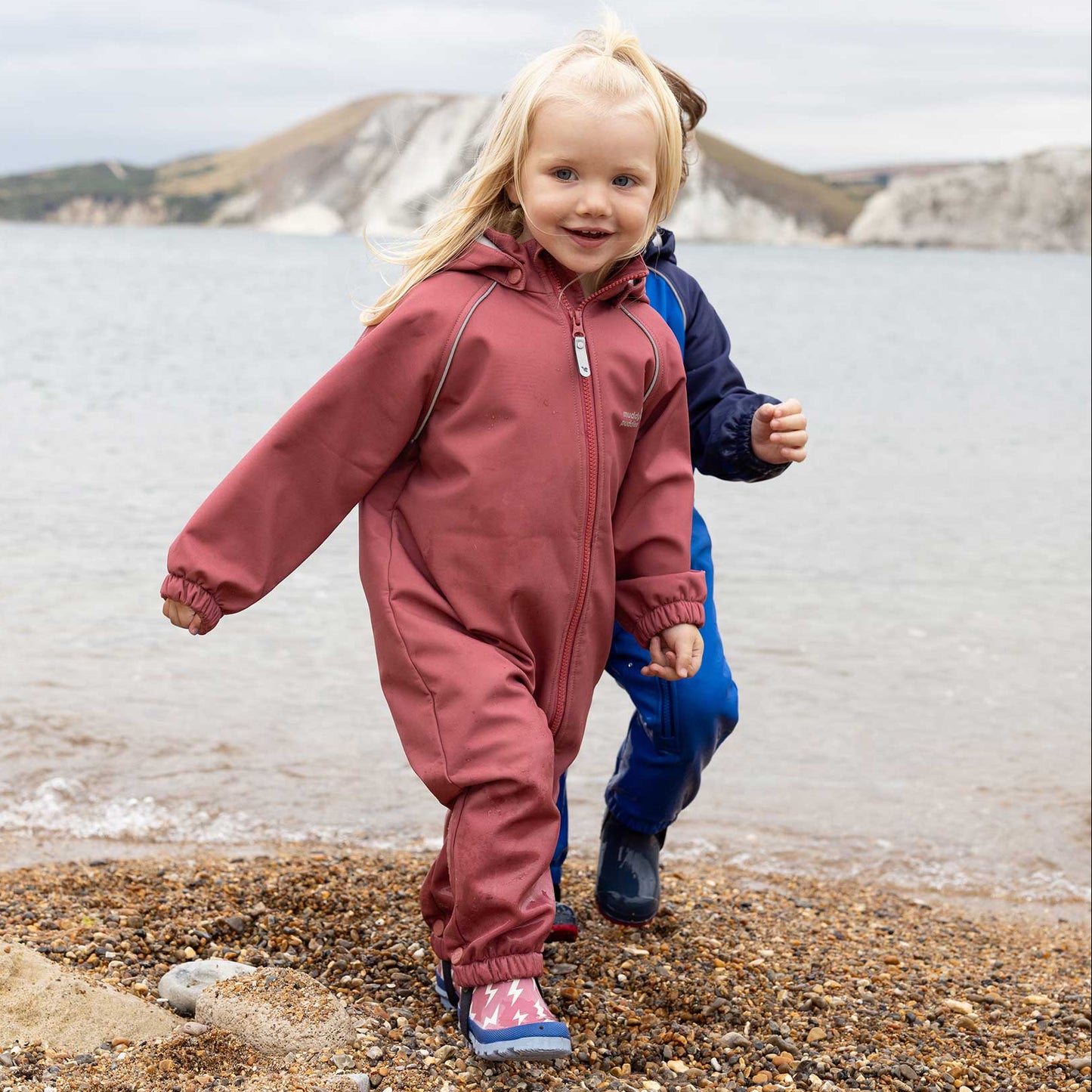 Child in durable Waterproof Softshell Puddle Suit Pink, smiling near a lake, showcasing weatherproof features ideal for outdoor adventures. Another child in blue suit in background.