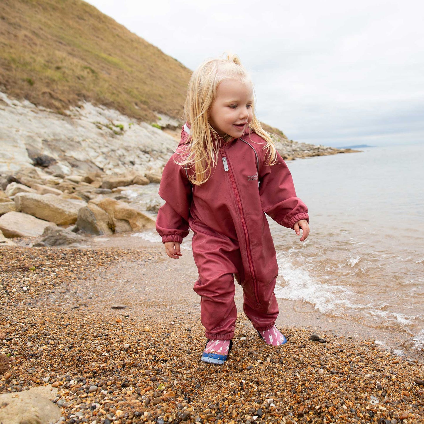 Toddler in durable Waterproof Softshell Puddle Suit Pink, exploring a pebbly shoreline, showcasing its waterproof, flexible design ideal for adventurous outdoor play.