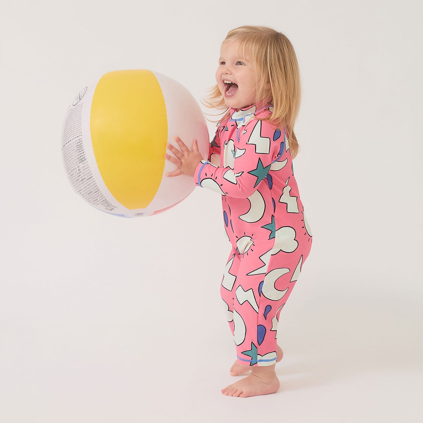 Toddler in UV Protective Surf Suit Pink Weather, holding a large beach ball. The suit offers UPF 50+ sun protection, perfect for outdoor adventures.